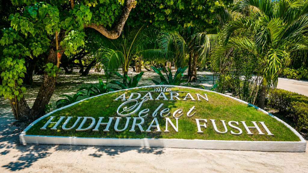Vstupní logo resortu Adaaran Select Hudhuran Fushi obklopené tropickou zelení na Maledivách.